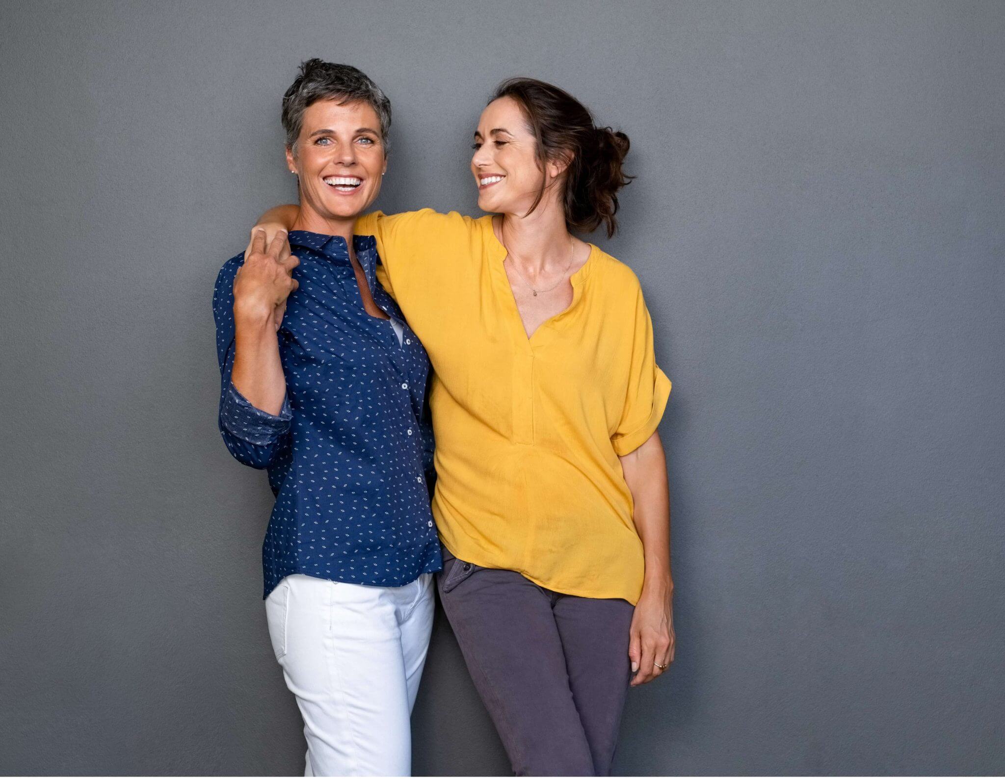 two women with their arms around each other. One is smiling at the other, the other is smiling at the camera.