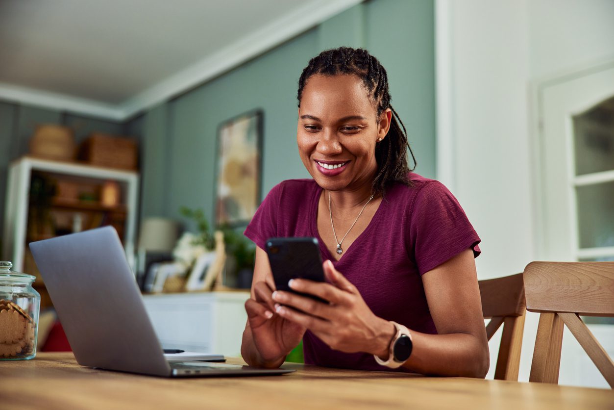 A smiling African-American woman texts messages on her phone while working on her laptop.
