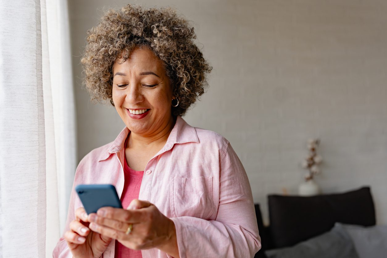 Middle-aged woman with curly hair wearing a pink shirt and looking at her iphone smiling.